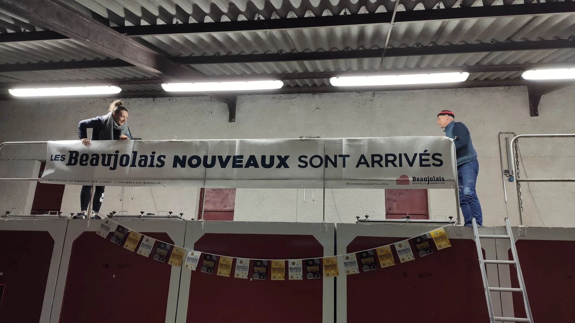 Deux personnes accrochent une bannière avec le texte « Les Beaujolais nouveaux sont arrivés » dans un bâtiment industriel.