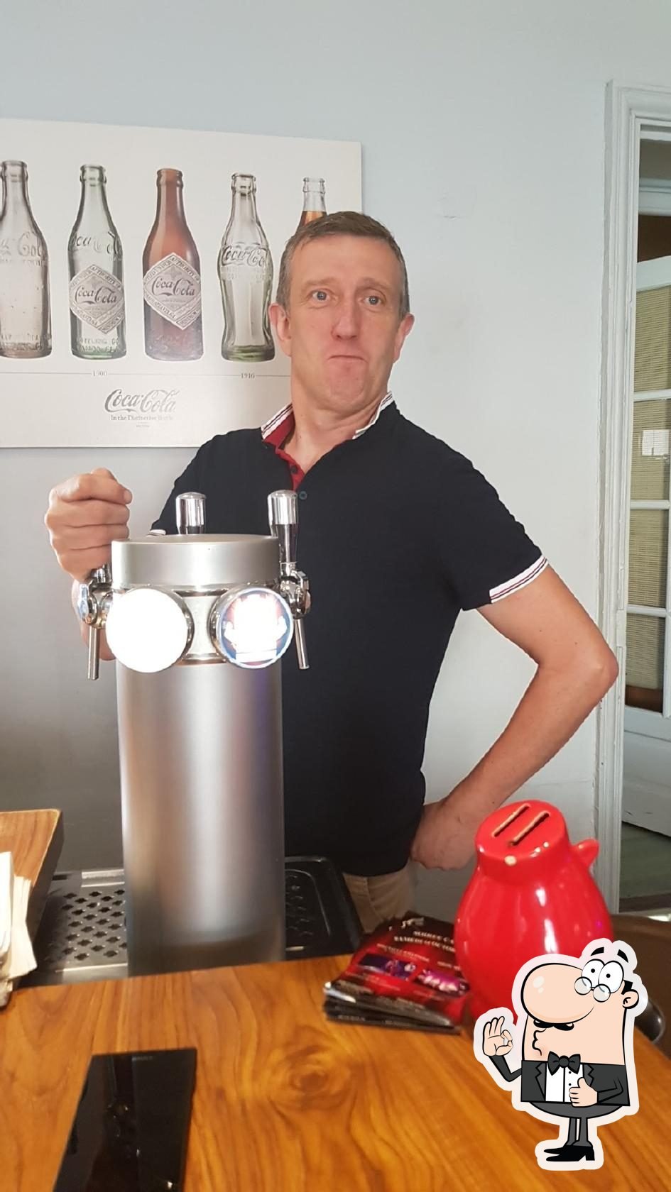 Un homme se tient derrière un comptoir, tenant une tireuse à bière, avec un tableau représentant différentes bouteilles de Coca-Cola en arrière-plan.