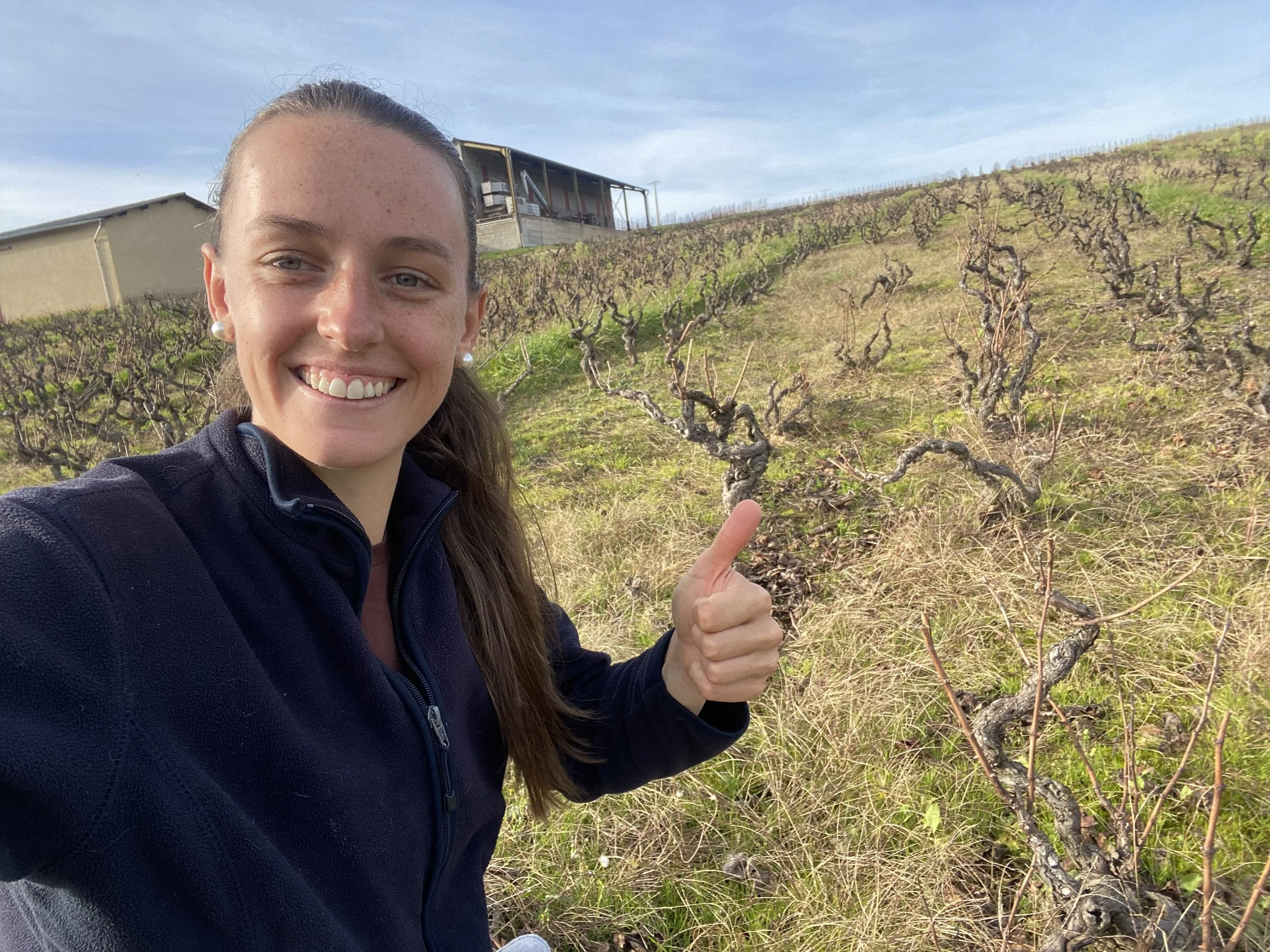 Une femme souriante fait un selfie dans un vignoble avec des vignes en hiver ou début de printemps, un bâtiment au fond.