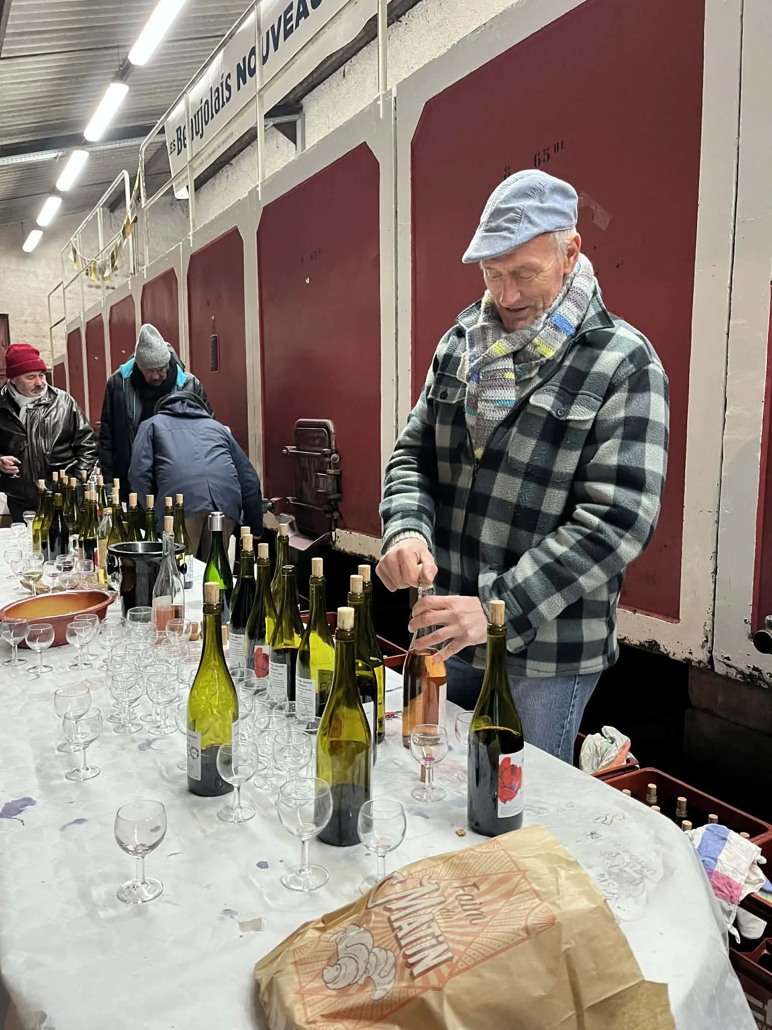 Un homme âgé avec une veste à carreaux, un béret gris et une écharpe colorée, en train de déguster ou de verser un vin dans un verre à l'aide d'un tire-bouchon, devant une table couverte de plusieurs bouteilles de vin et de petits verres à vin dans une cave ou une salle de dégustation.