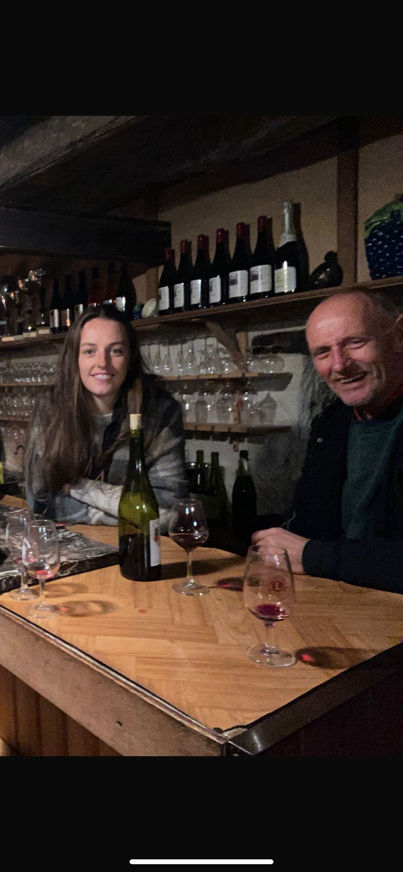 Deux personnes assises à un bar en train de sourire, entourées de bouteilles de vin et de verres à vin sur le comptoir en bois.