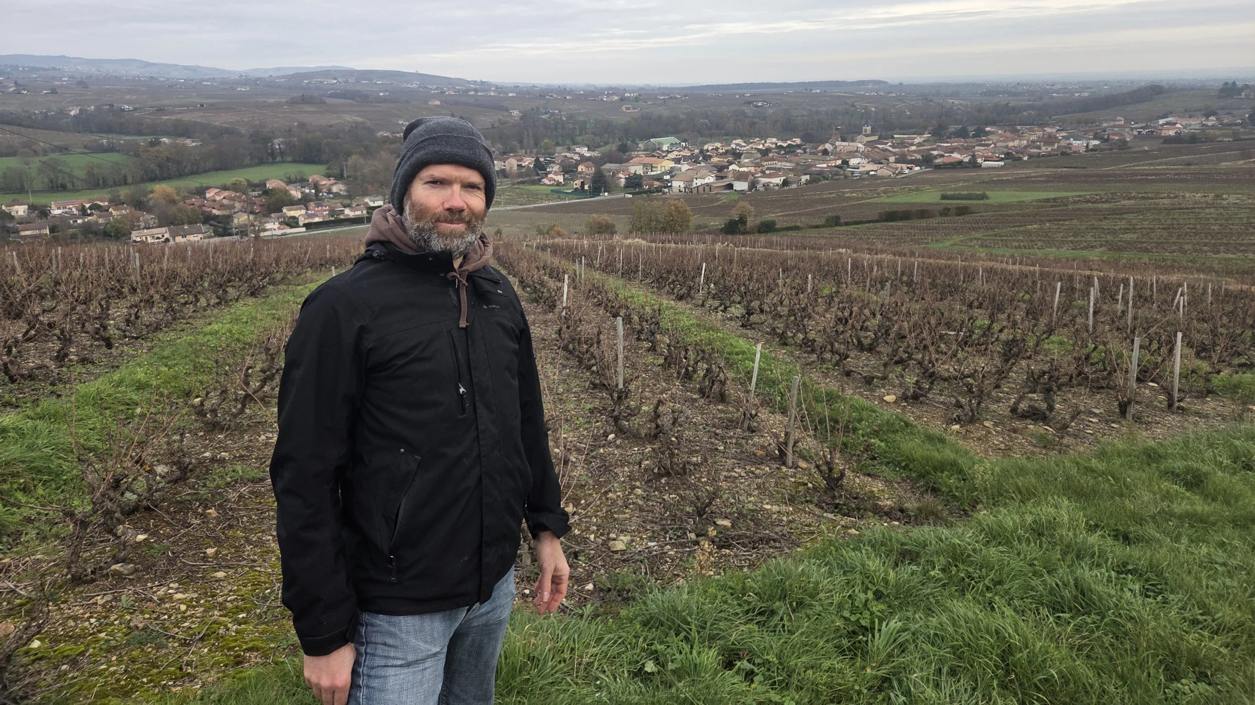Un homme avec un manteau noir, un bonnet gris et un jean se tient dans un vignoble, avec un village et des collines en arrière-plan, par un ciel nuageux.