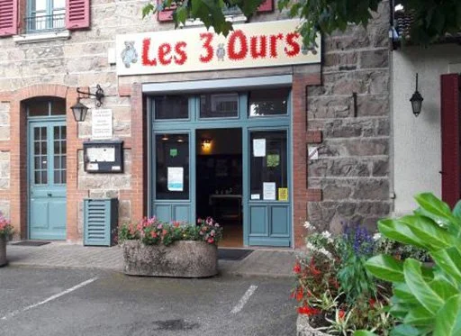 Façade d'un restaurant avec une enseigne 'Les 3 Ours', entrée en bois foncé, fleurs décoratives devant, lampes murales, et fenêtres en verre.