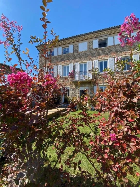 Maison en pierre avec fenêtres à volets blancs, entourée de plantes et de fleurs roses dans un jardin sous un ciel bleu clair.