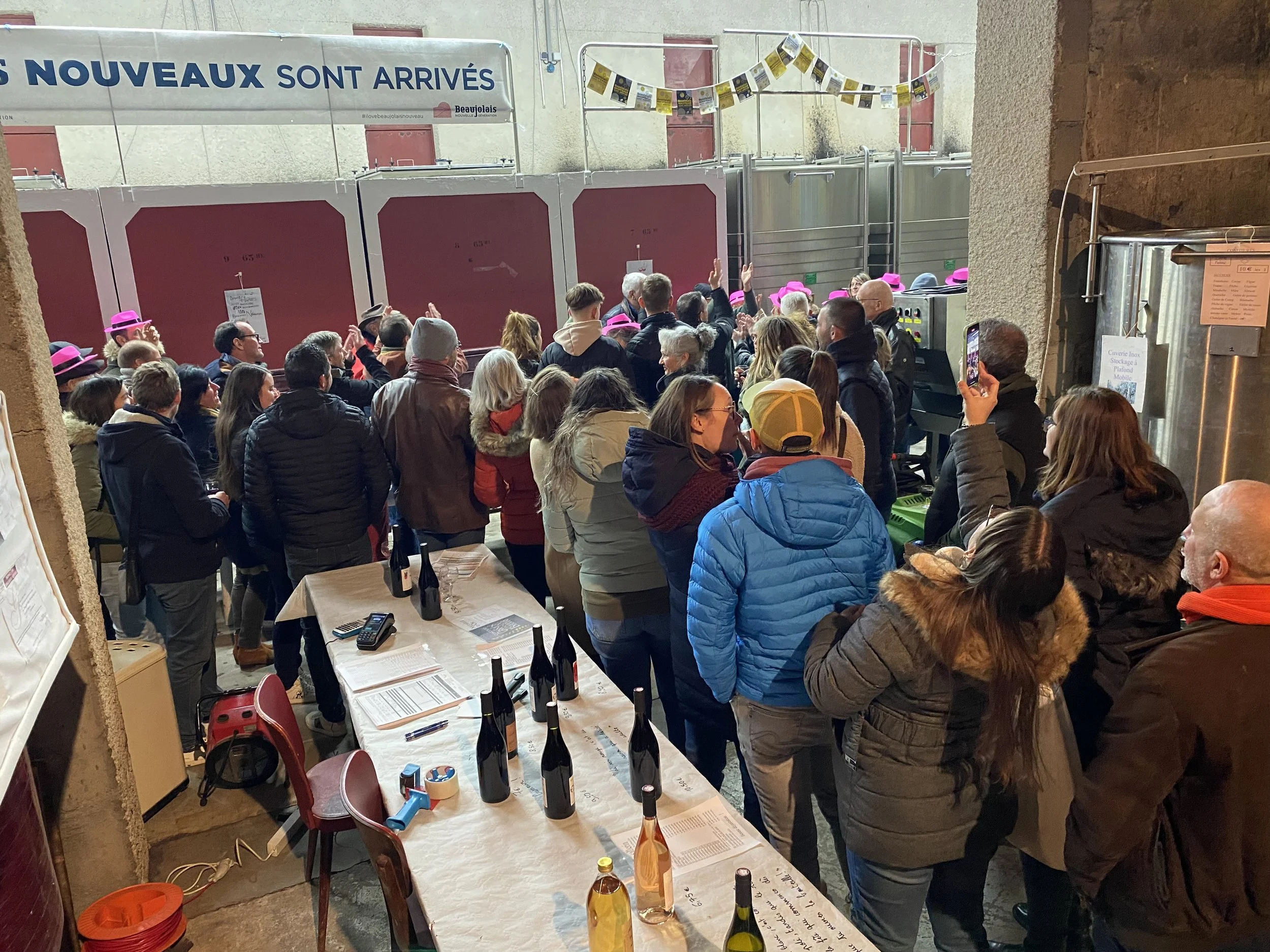 Foule de personnes faisant la queue dans un endroit couvert, probablement une cave ou une salle de dégustation de vins, avec plusieurs bouteilles sur une table.