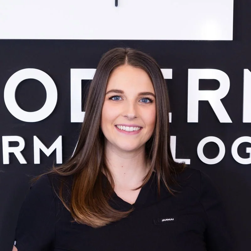 Young woman with long brown hair, smiling, standing in front of a black and white sign.