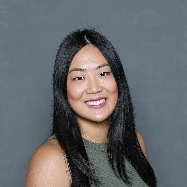 A young Asian woman with long black hair and a smile, wearing a sleeveless green top, posing against a gray background.