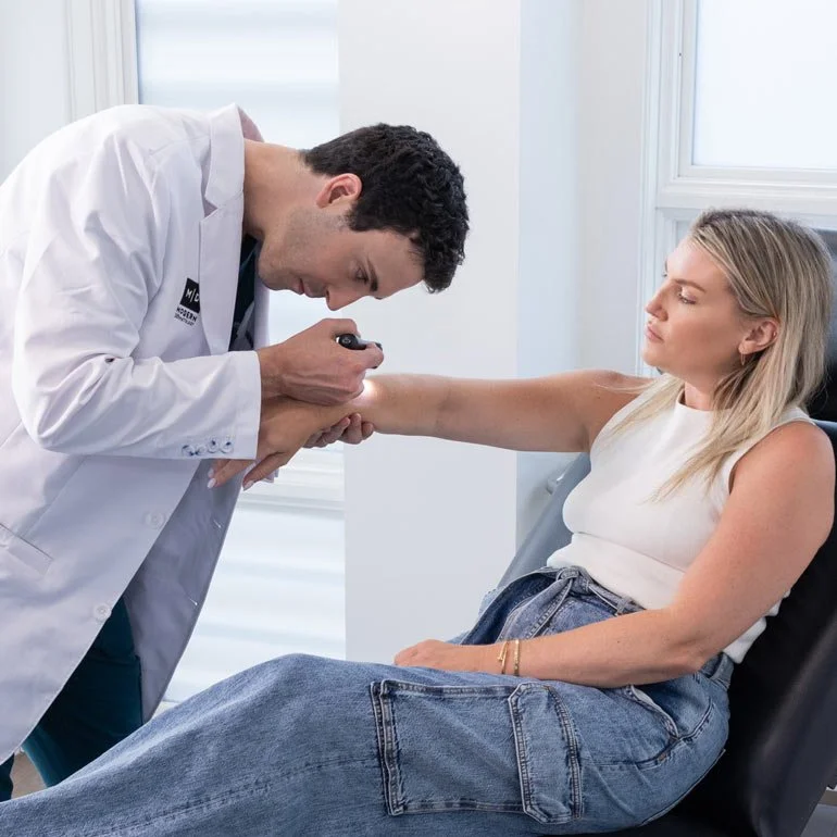 A healthcare professional wearing a white coat is examining a woman's arm with a medical instrument in a clinical setting, with natural light coming through a window.