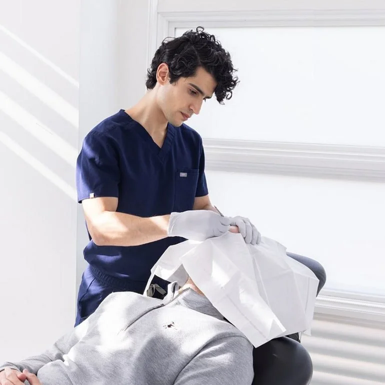 A healthcare professional in navy scrubs and white gloves preparing to perform a procedure on a patient lying in a medical setting.
