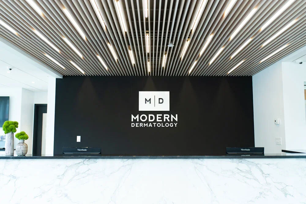 Interior of a modern dermatology clinic with a black wall displaying the logo 'MD Modern Dermatology' in white text, with a marble reception desk and decorative green plants on the left.