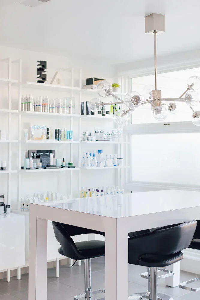 Modern skincare clinic with white shelves filled with skincare products, black chairs, white table, and a contemporary chandelier in a bright room.