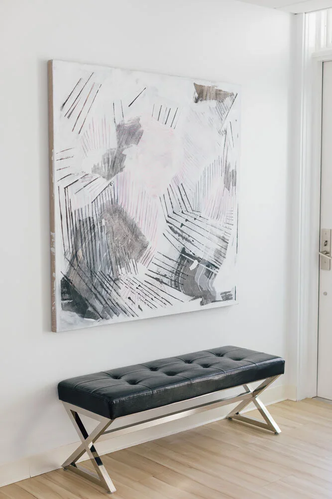 Modern black leather bench with metal X-shaped legs in front of abstract black and white artwork on white wall in a bright room.