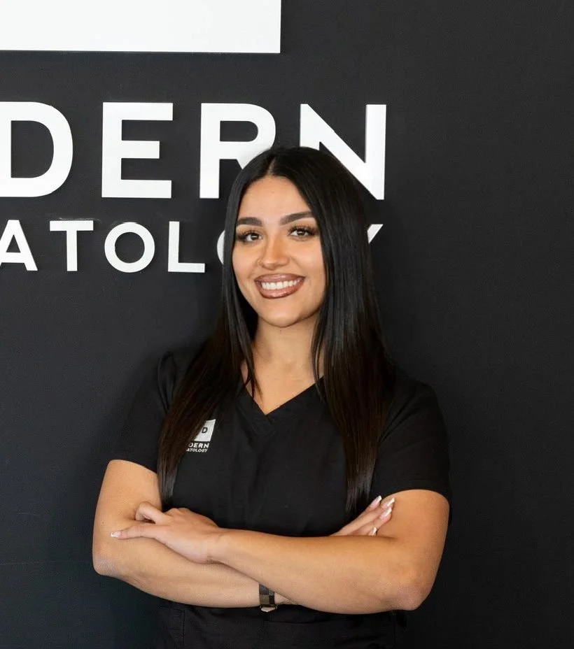 A woman with long black hair, smiling and crossing her arms, standing in front of a black wall with white text that reads 'DERN ATOL'.