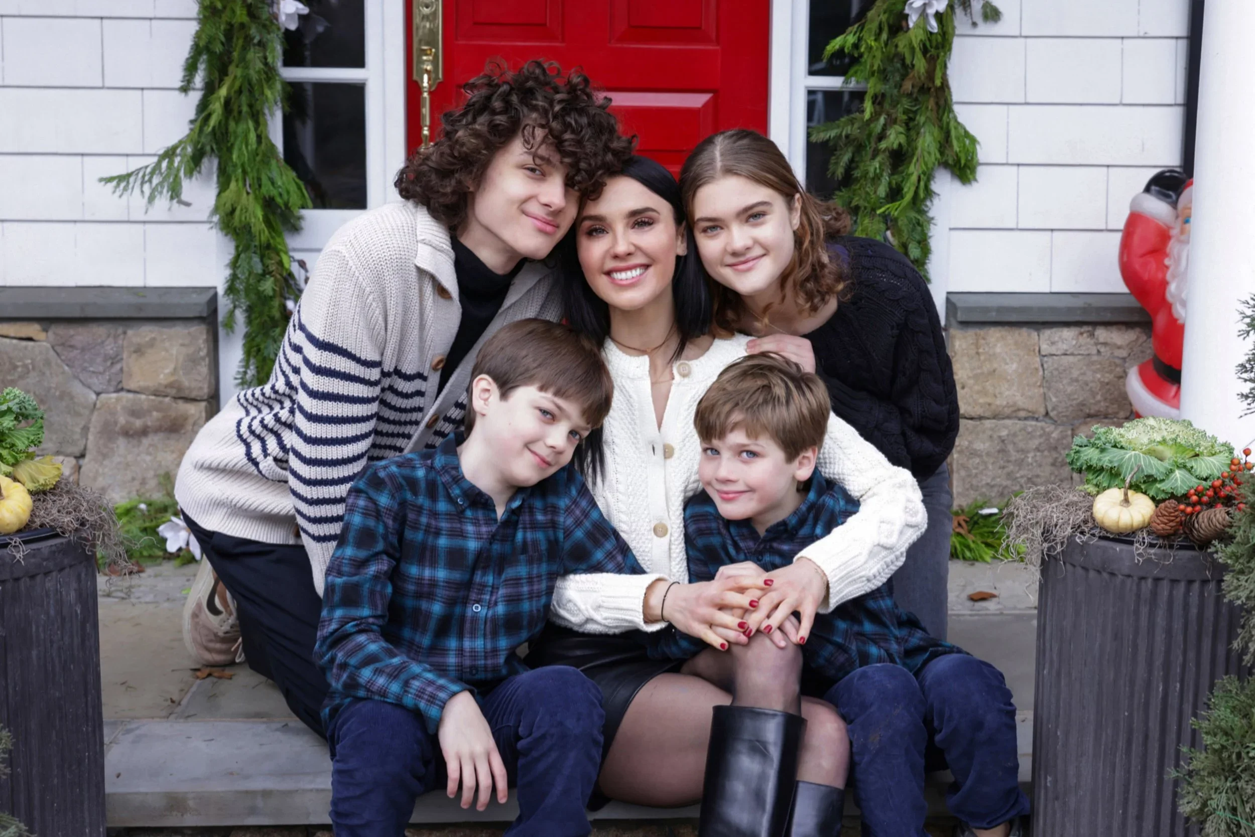 A woman and five children sitting on a front porch in front of a white house with a red door, decorated with Christmas greenery and a Santa Claus figure to the side, smiling and posing for a photo.