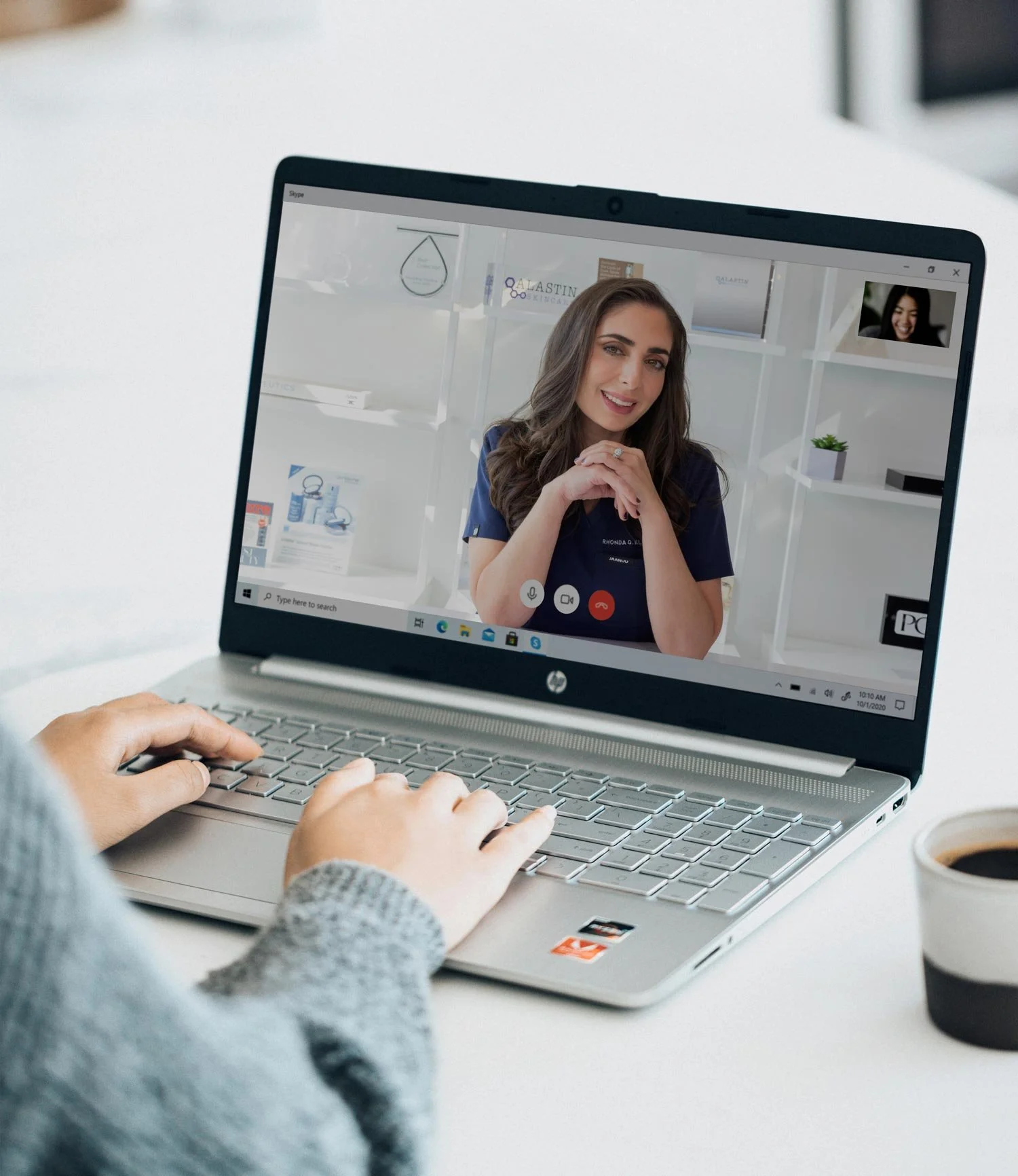 Laptop screen showing a woman smiling during a video call with another woman with long dark hair, in a bright room with white shelves and office decor.