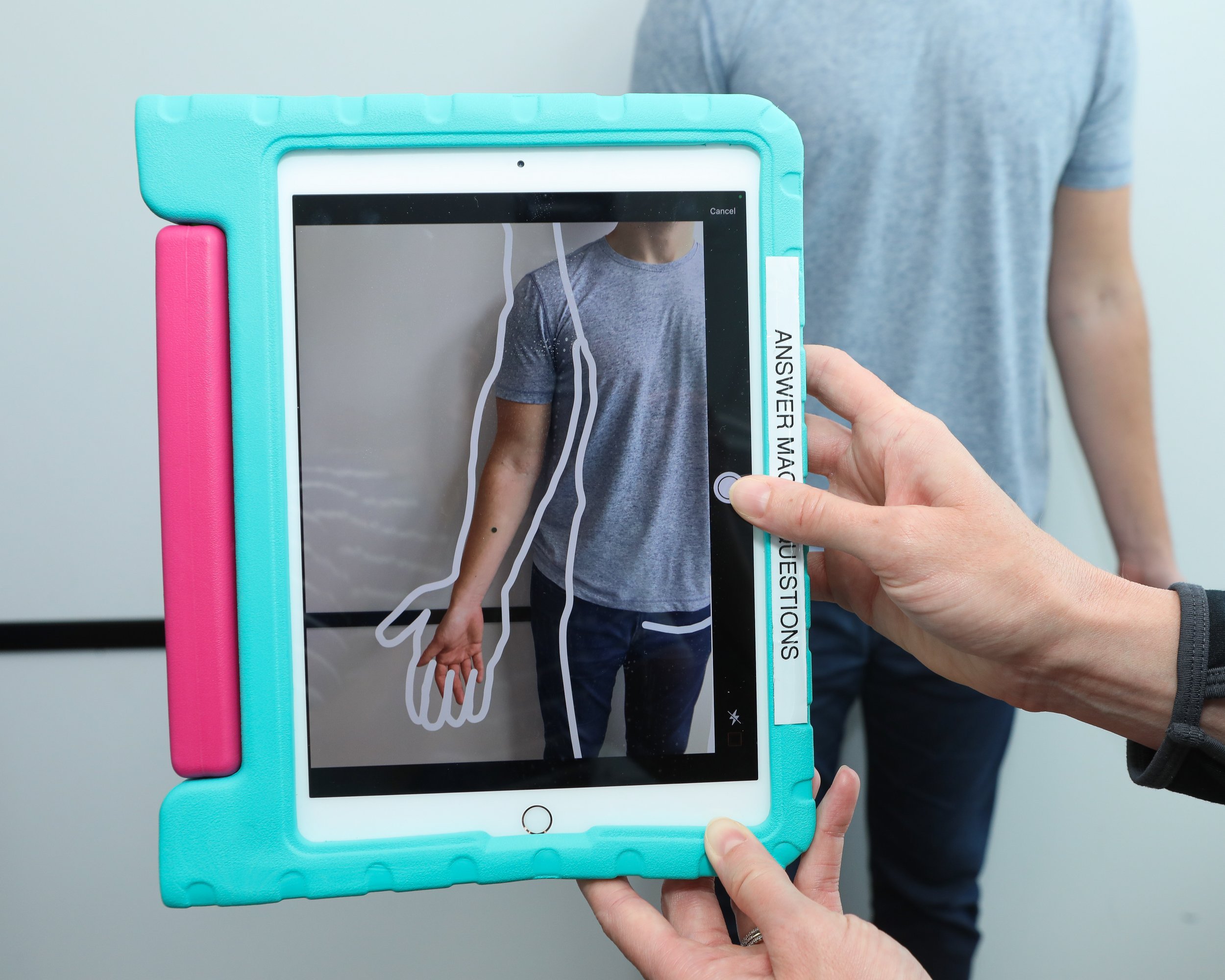 A person holding an iPad with a colorful protective case, using an app that labels a diagram of a human body. The person is wearing a gray t-shirt.