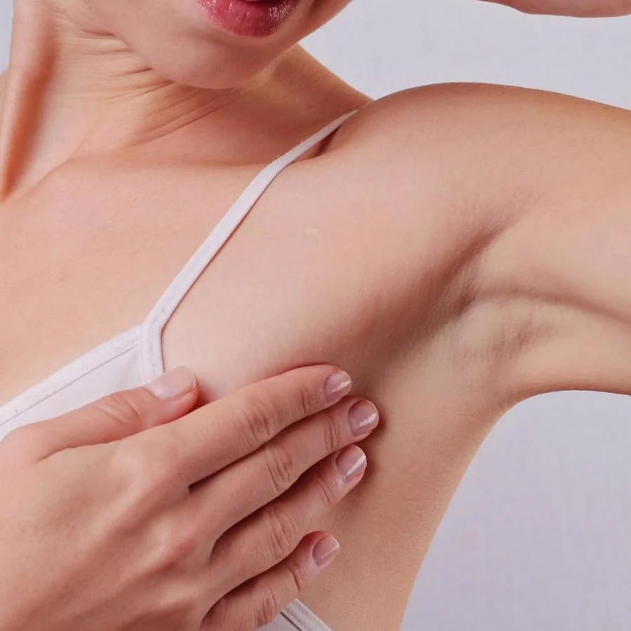 A person with fair skin wearing a white tank top is raising their arm, exposing their armpit, and touching it with their hand.