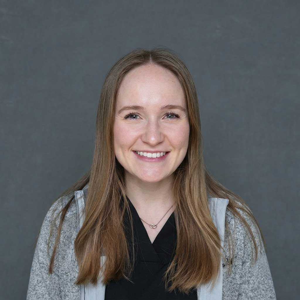 A young woman with long, light brown hair, smiling, standing outdoors in front of a white, horizontal wooden-paneled wall.