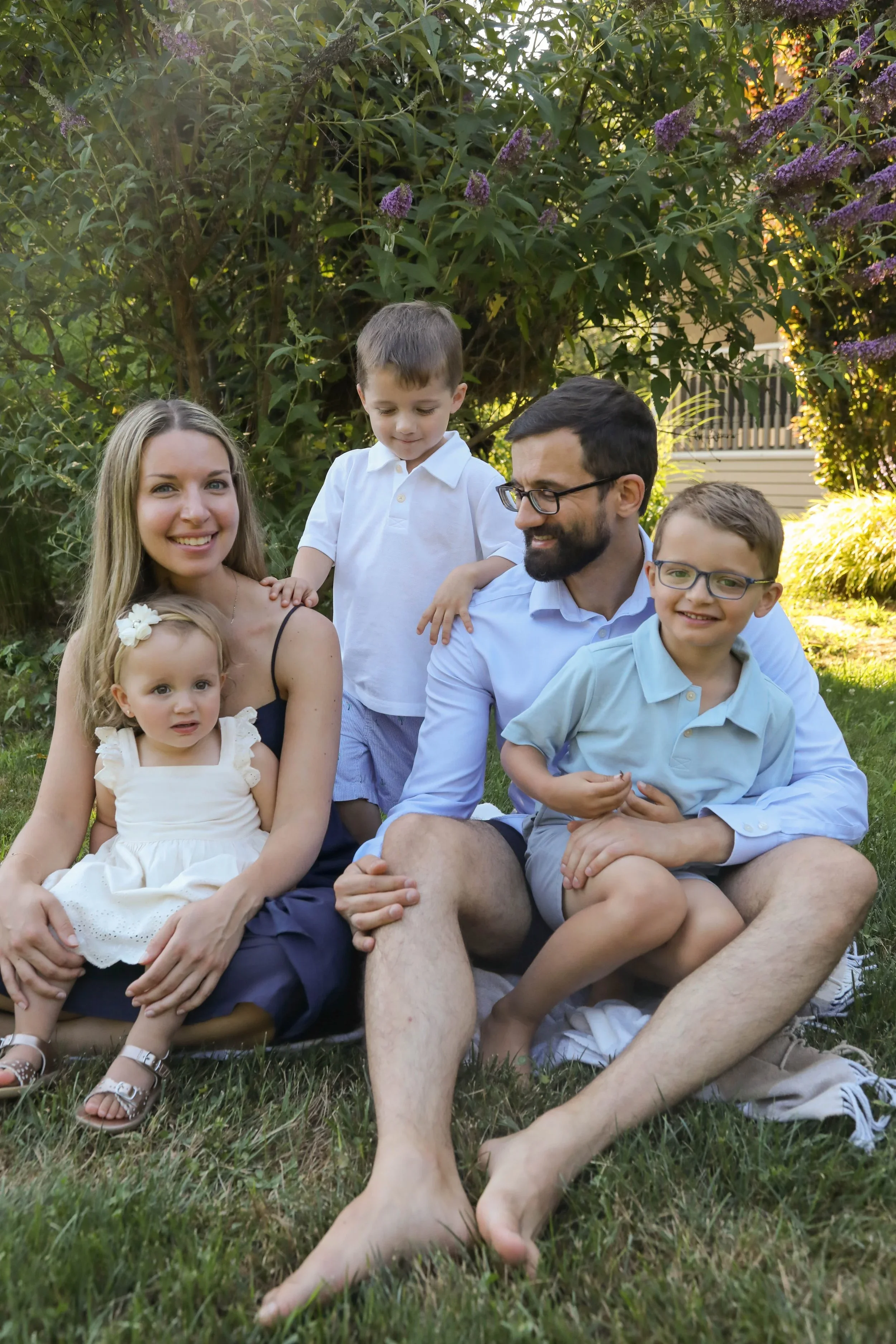 Family of six sitting and lying on grass in a garden, smiling, surrounded by green plants and trees.