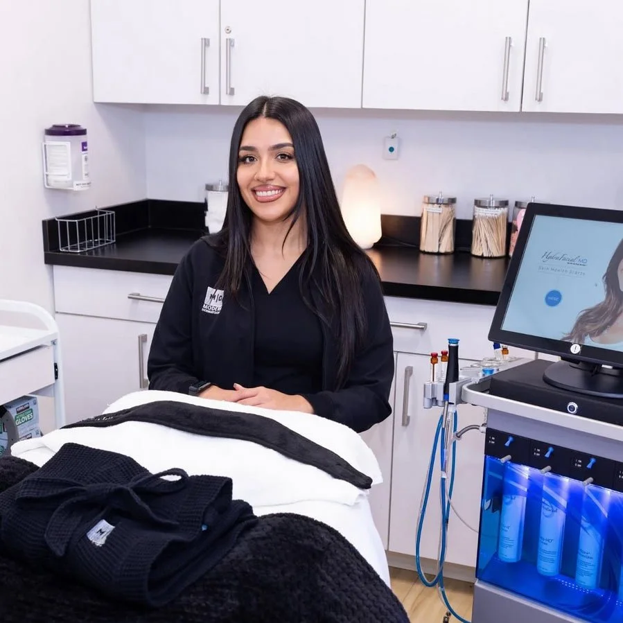 A smiling woman with long dark hair in a medical or spa treatment room, standing behind a treatment bed with black clothing on it. The room has white cabinets, a computer monitor, and various medical supplies.