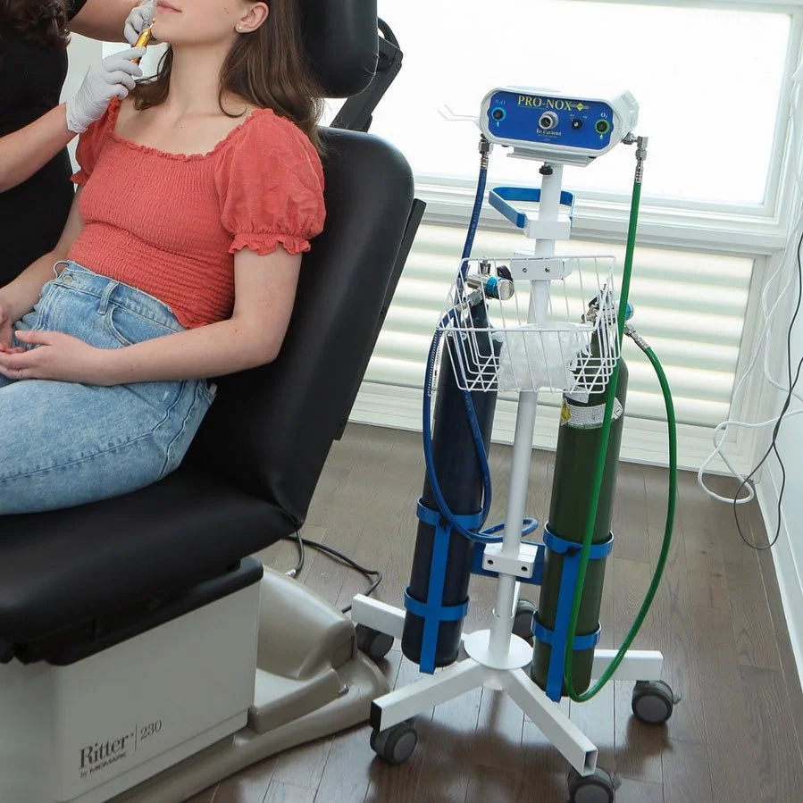 A woman sitting in a medical chair receiving a treatment, with a healthcare professional administering an injection or local anesthesia in her face. Next to her, there is medical equipment on a wheeled stand, including a oxygen tank and a device labeled PRO-NOX for pain relief during dental procedures.