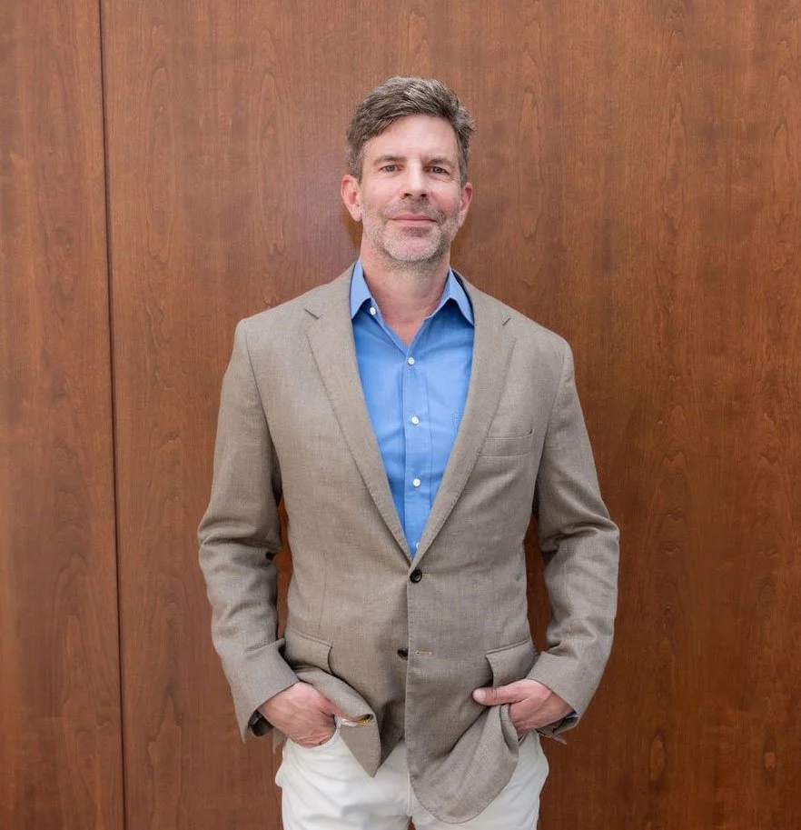 A middle-aged man in a beige suit with a blue dress shirt, standing in front of a wooden wall, with his hands in his pockets, smiling slightly.