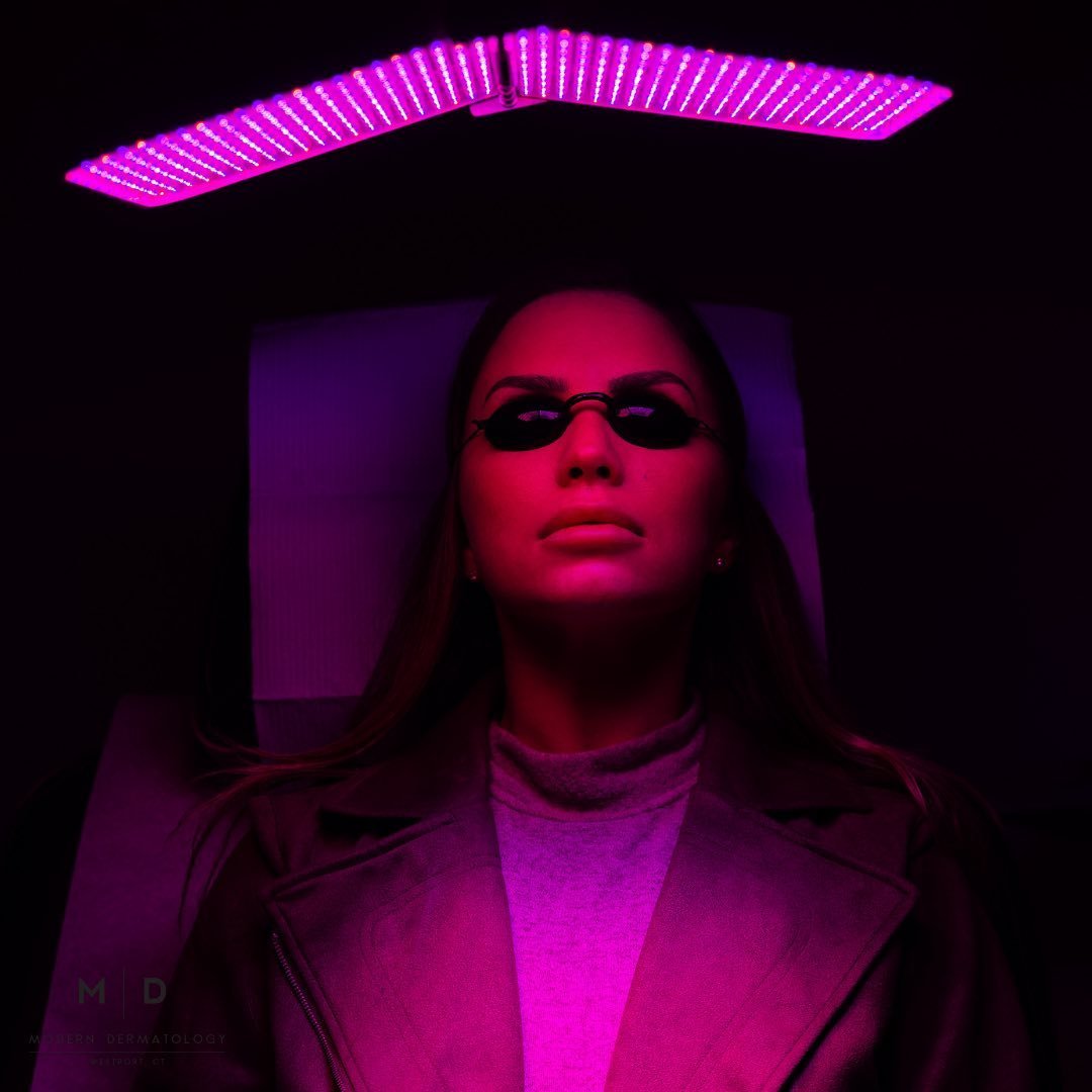 A woman wearing black oval sunglasses and a leather jacket, illuminated by pink and purple lighting, lying on a bed with a pink light fixture above her head.