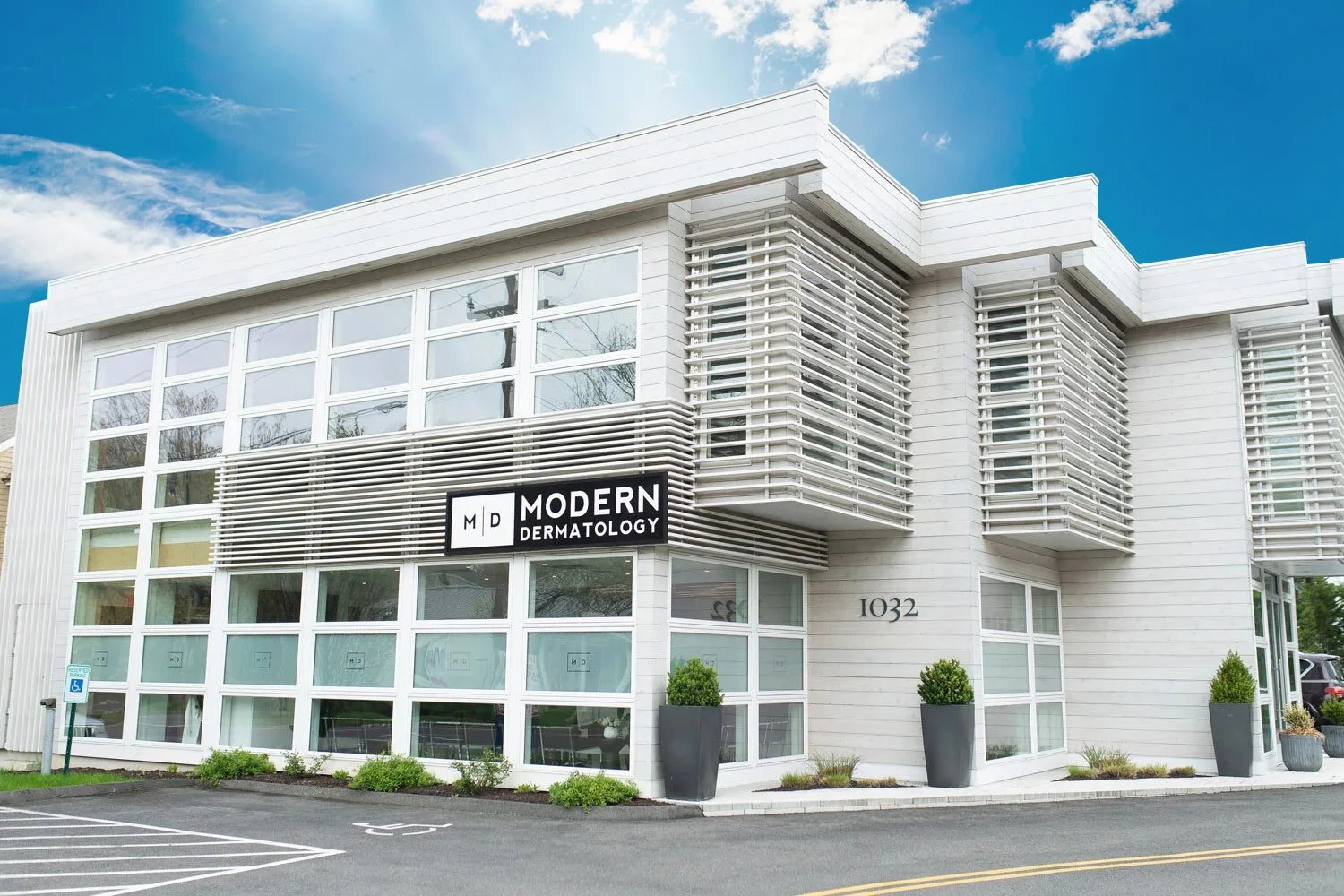 Modern dermatologist office building with white exterior, large glass windows, and a black sign that reads "Modern Dermatology." The building number "1032" is visible. There are large potted plants outside and a parking lot nearby, under a partly cloudy sky.