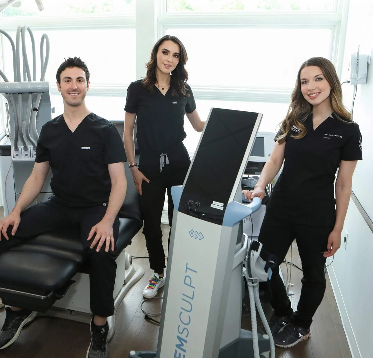 Three healthcare professionals in black scrubs inside a medical office with equipment around, some smiling at the camera, one sitting on an examination table, another standing beside a medical device, and the third standing behind them near a computer in a well-lit room.