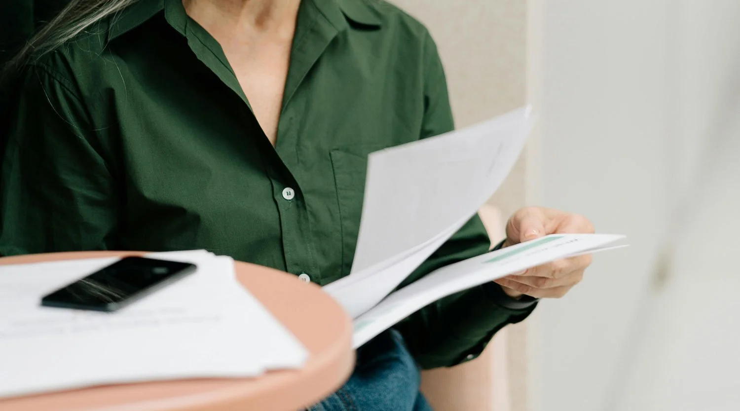 Person wearing a green shirt reading papers at a table, with a phone on a stack of documents.