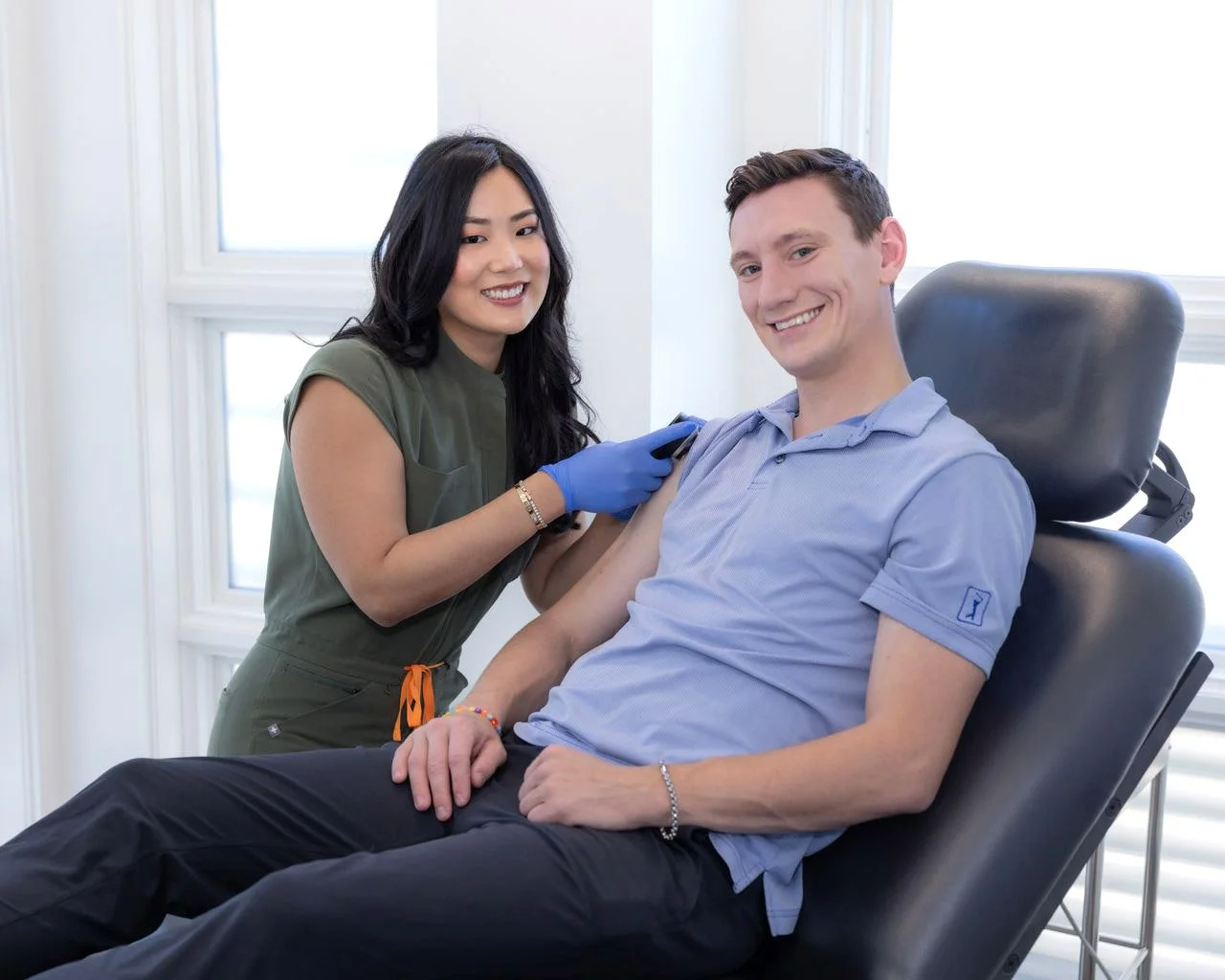 A woman in medical scrubs and blue gloves administers a vaccine to a smiling man seated in a recline chair. The setting is a bright room with white walls and large windows.