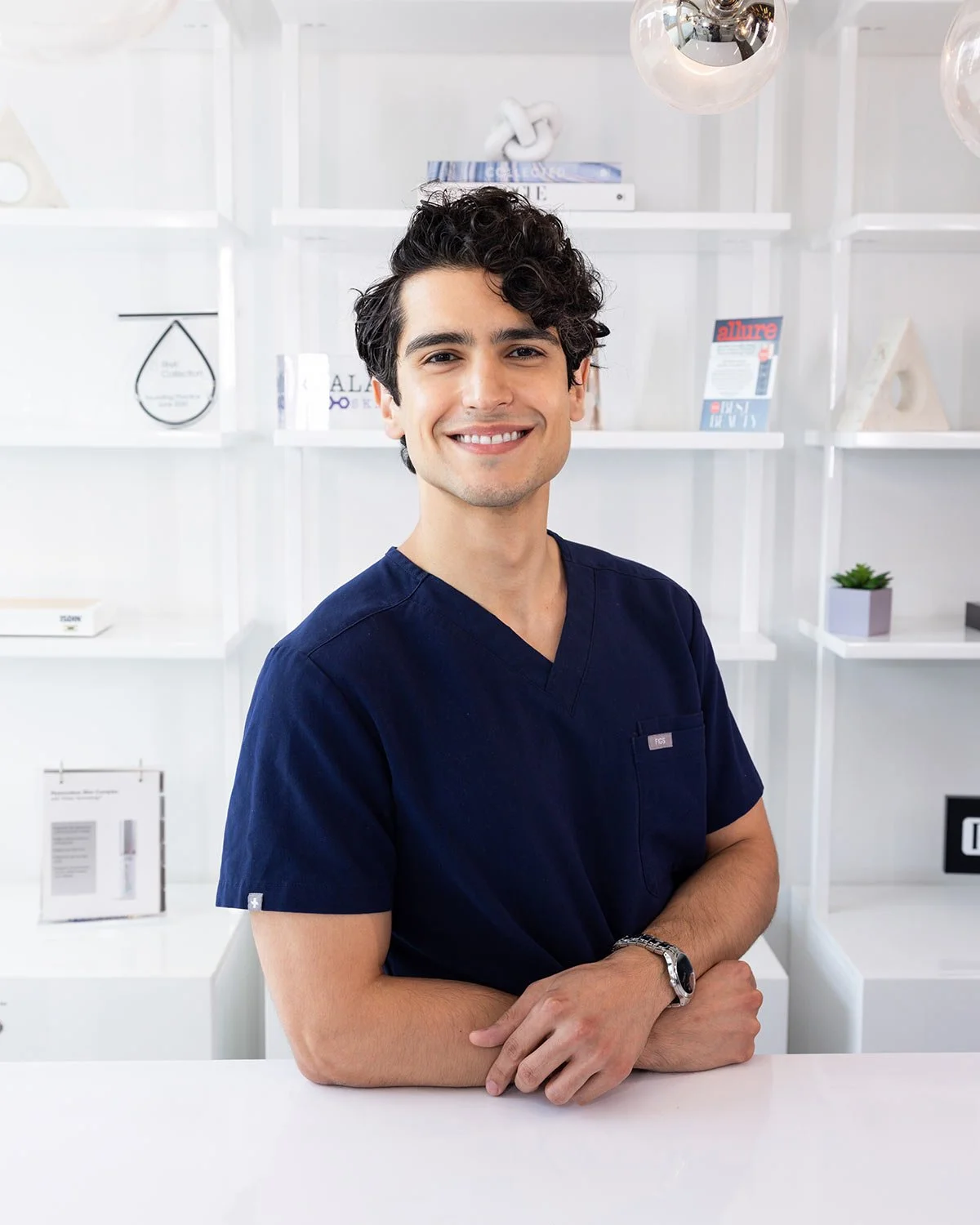 A smiling young man with dark curly hair wearing navy scrubs, sitting at a white table in a modern, minimalistic office or clinic setting with white shelves and decorative items in the background.