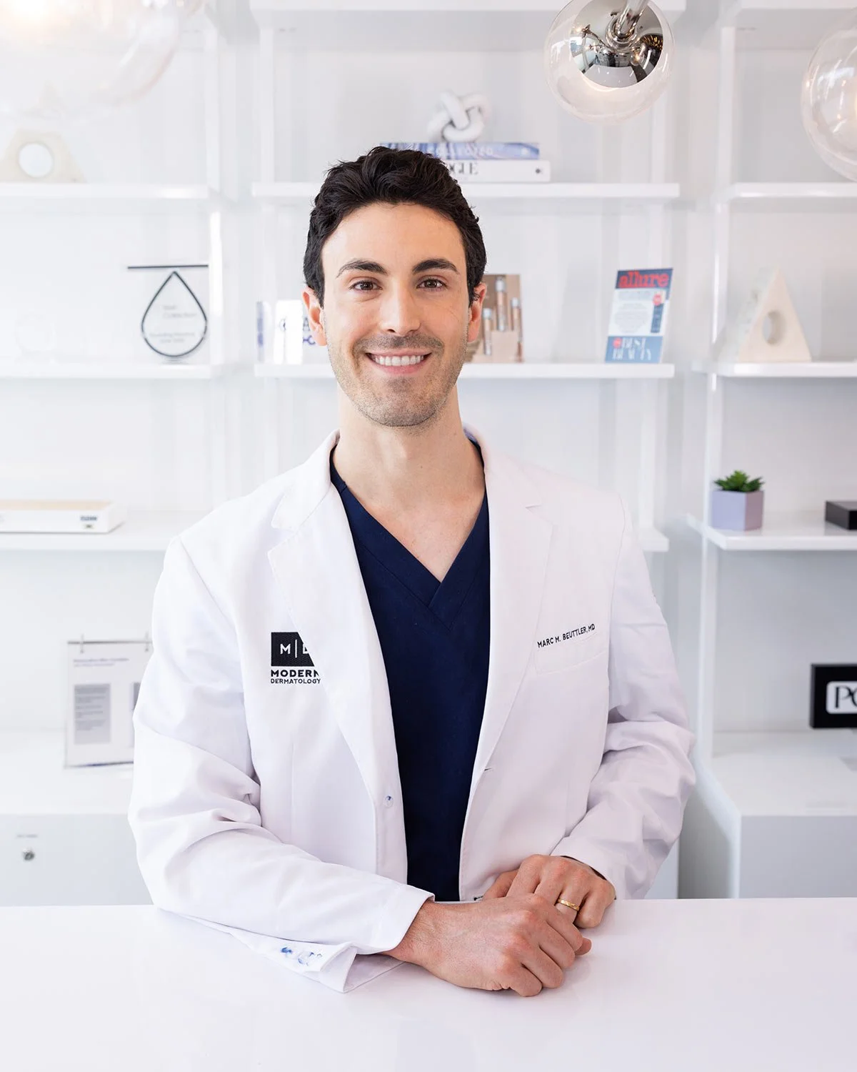 A smiling male doctor in a white coat standing in a modern medical office with white shelves and decor in the background.