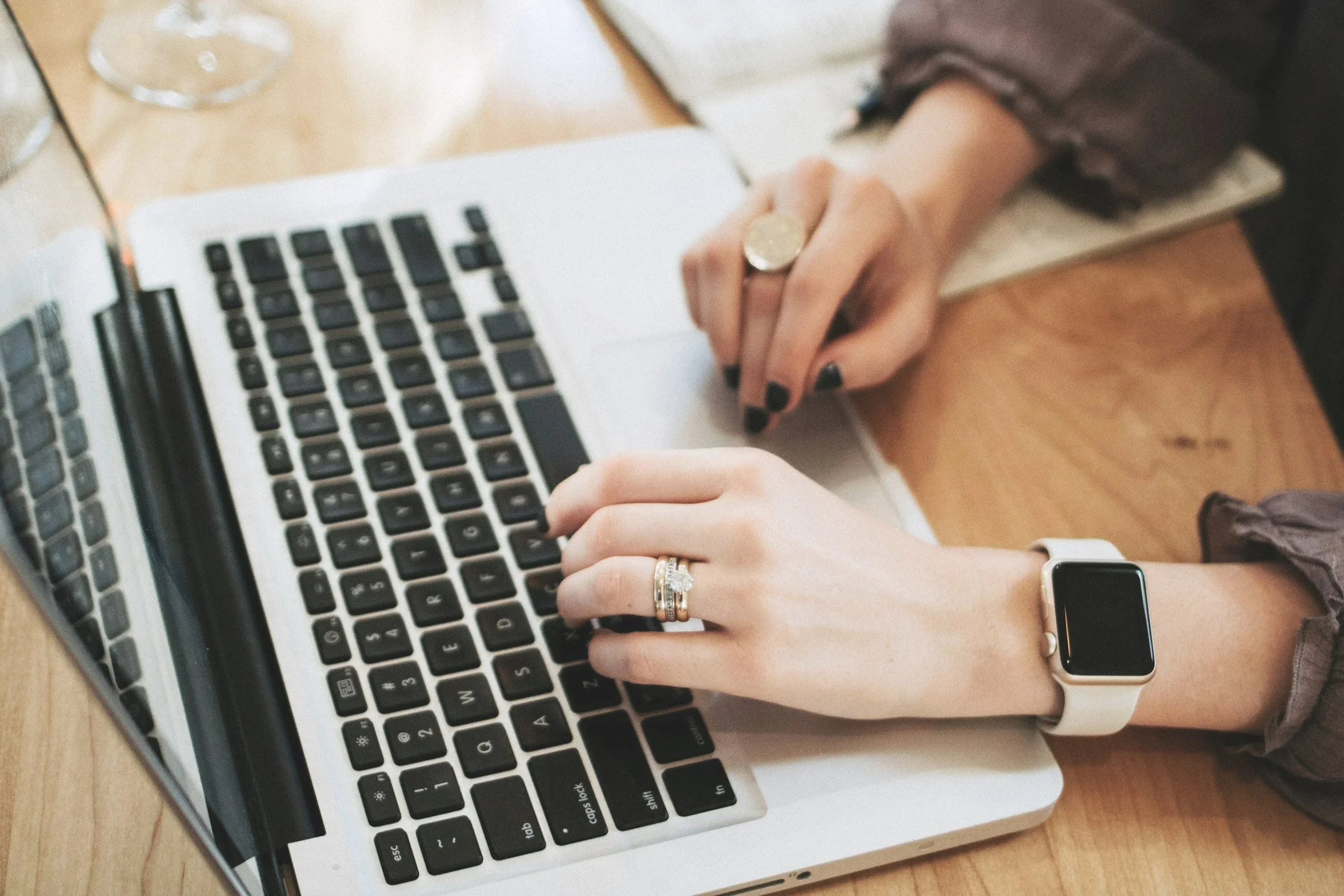 Two people working on a laptop at a table, one wearing a wedding ring and smartwatch, the other wearing a ring and black nail polish.