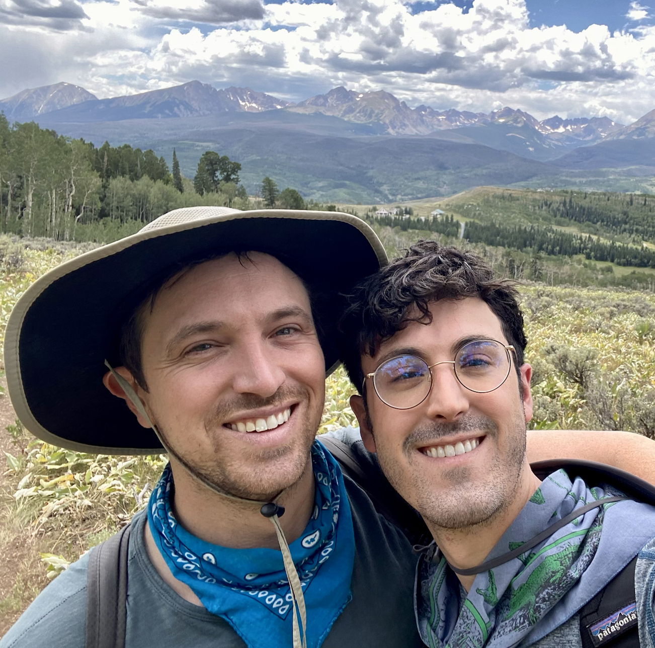 Two smiling men taking a selfie outdoors in a scenic mountainous landscape with trees, fields, and mountains in the background.
