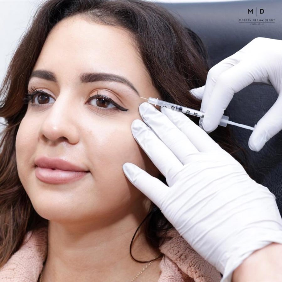 A woman receiving a cosmetic injection near her eye from a medical professional wearing white gloves.