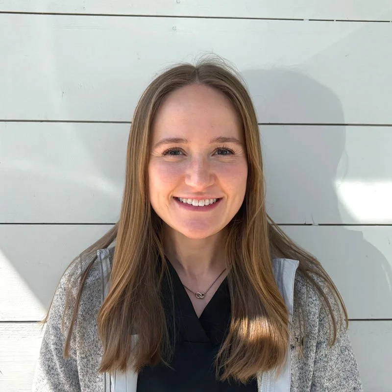A young woman with long, light brown hair, smiling, standing outdoors in front of a white, horizontal wooden-paneled wall.