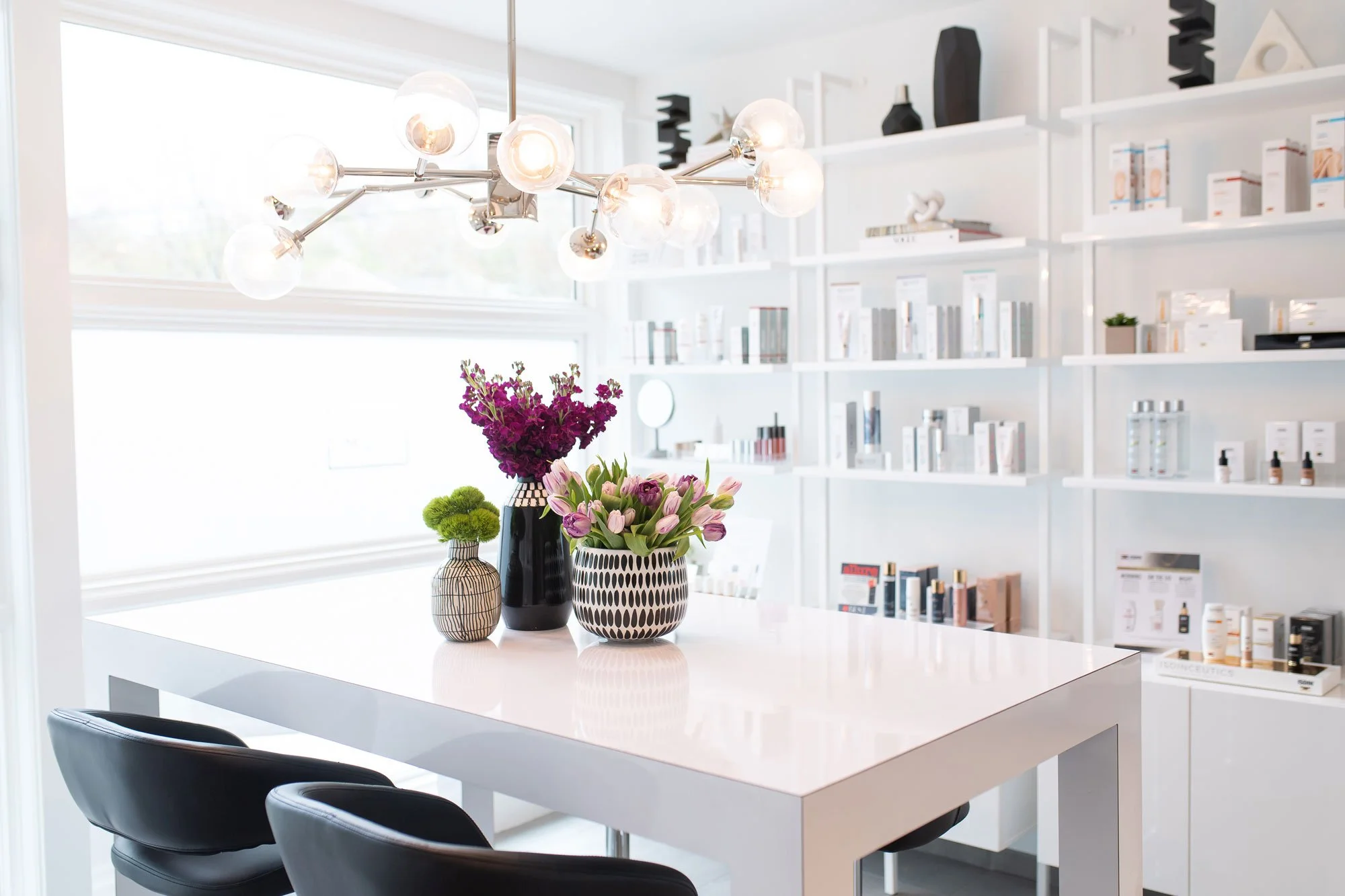 A modern white kitchen or dining area with a white table, black chairs, three vases with purple and pink flowers, a black, white, and beige patterned vase, a black pitcher, and a chandelier with multiple glass bulbs hanging from the ceiling. White shelves with various items in the background.