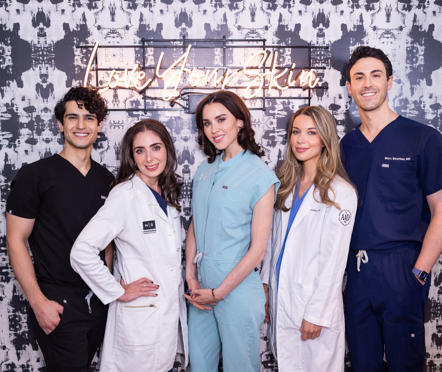 Medical professionals standing in front of a wall with a neon sign that reads "Love Your Skin"; they are wearing scrubs and lab coats.