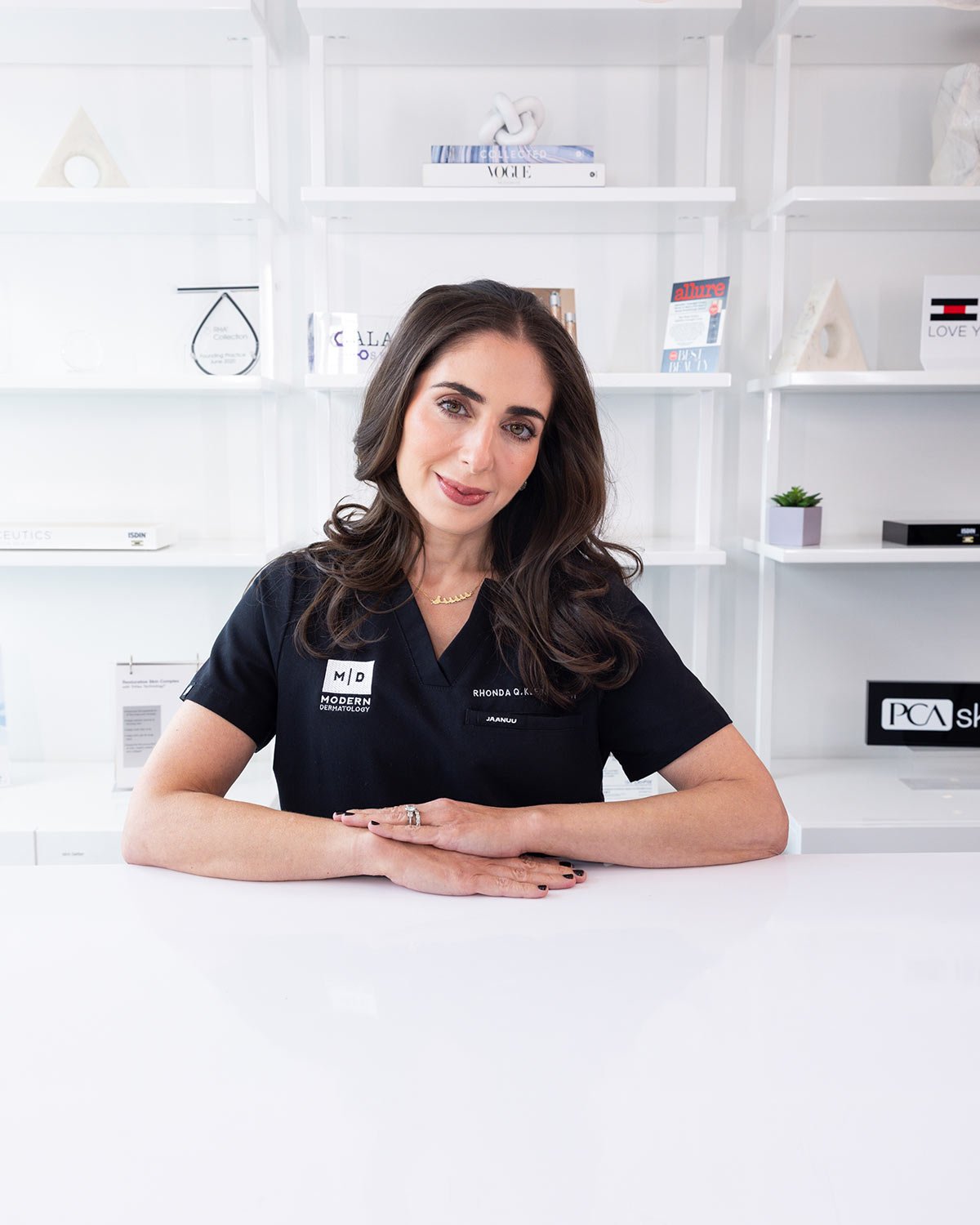 A woman with long dark hair sitting at a white table, wearing a navy medical scrub with a name tag, in a modern, minimalist office with white shelves and decor in the background.