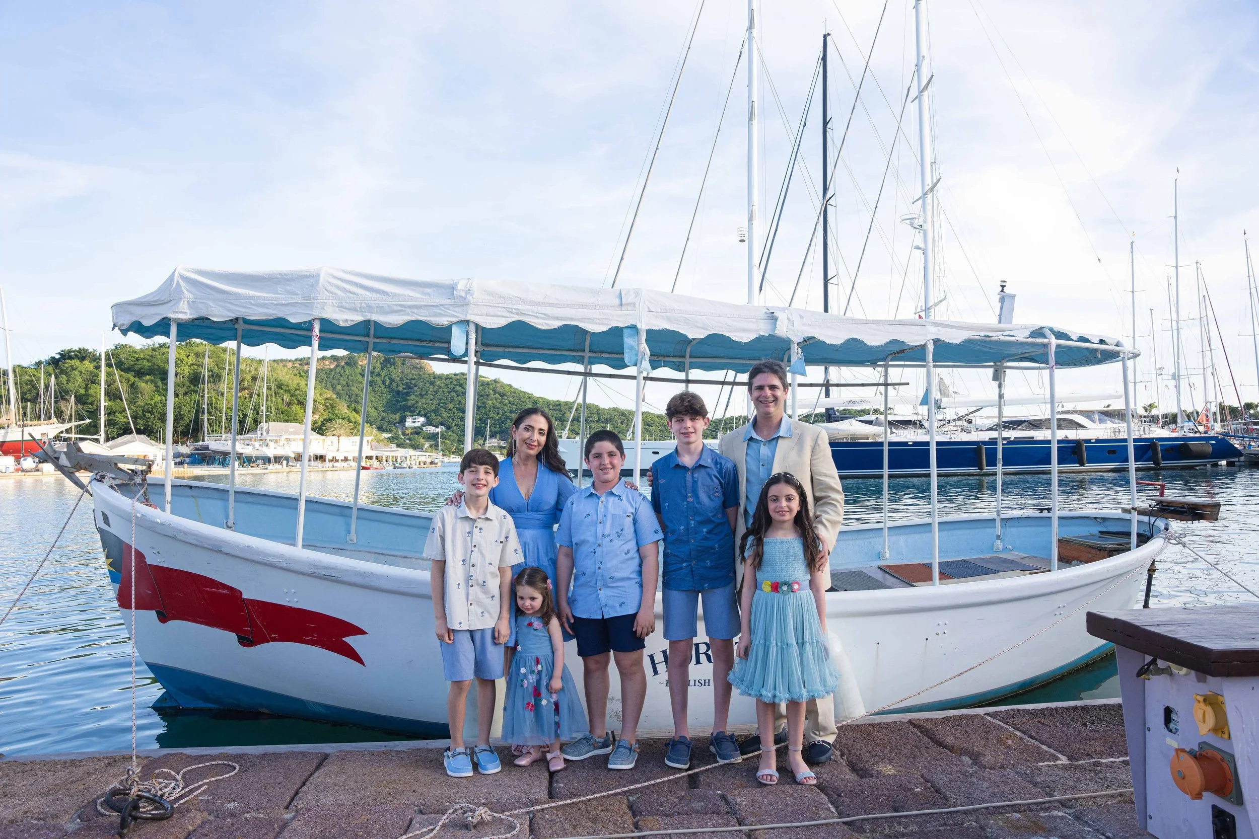 Group of seven people, including adults and children, standing in front of a boat docked at a marina with sailboats and trees in the background.
