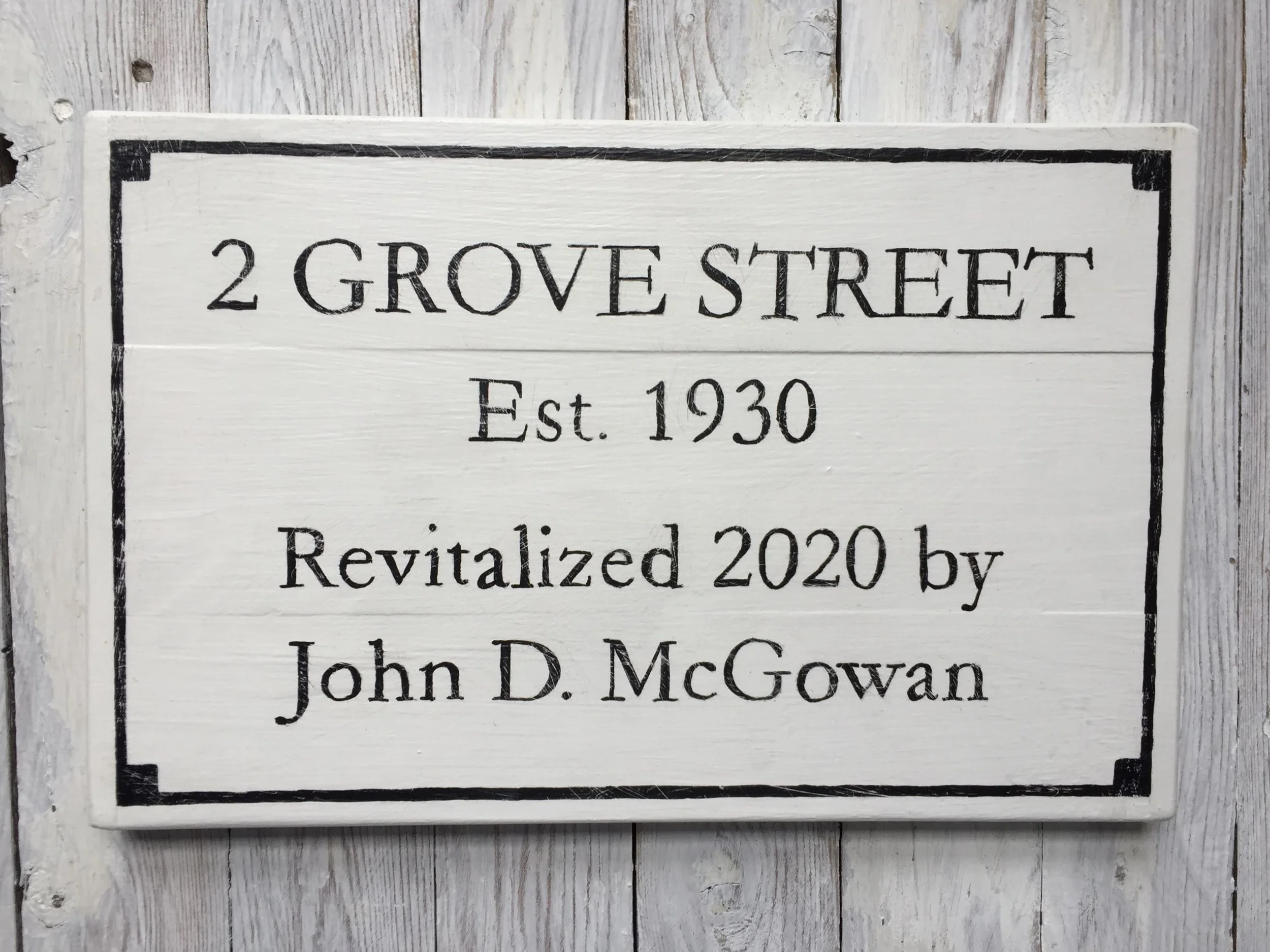 A decorative white wooden sign with black border and text, displaying the address '2 Grove Street,' established in 1930, revitalized in 2020 by John D. McGowan