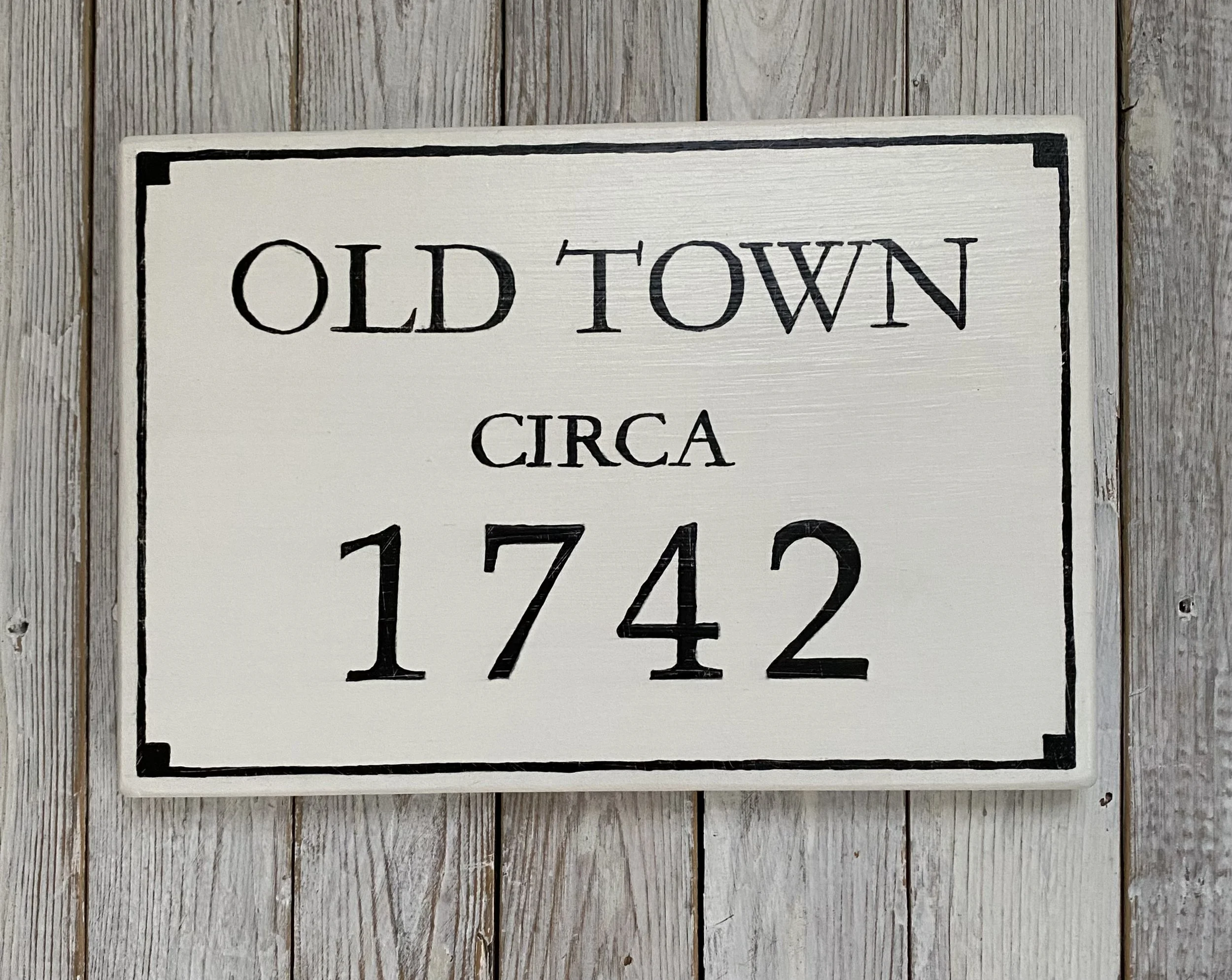 White wooden sign with black border and text reading "Old Town Circa 1742" against a weathered wooden wall.