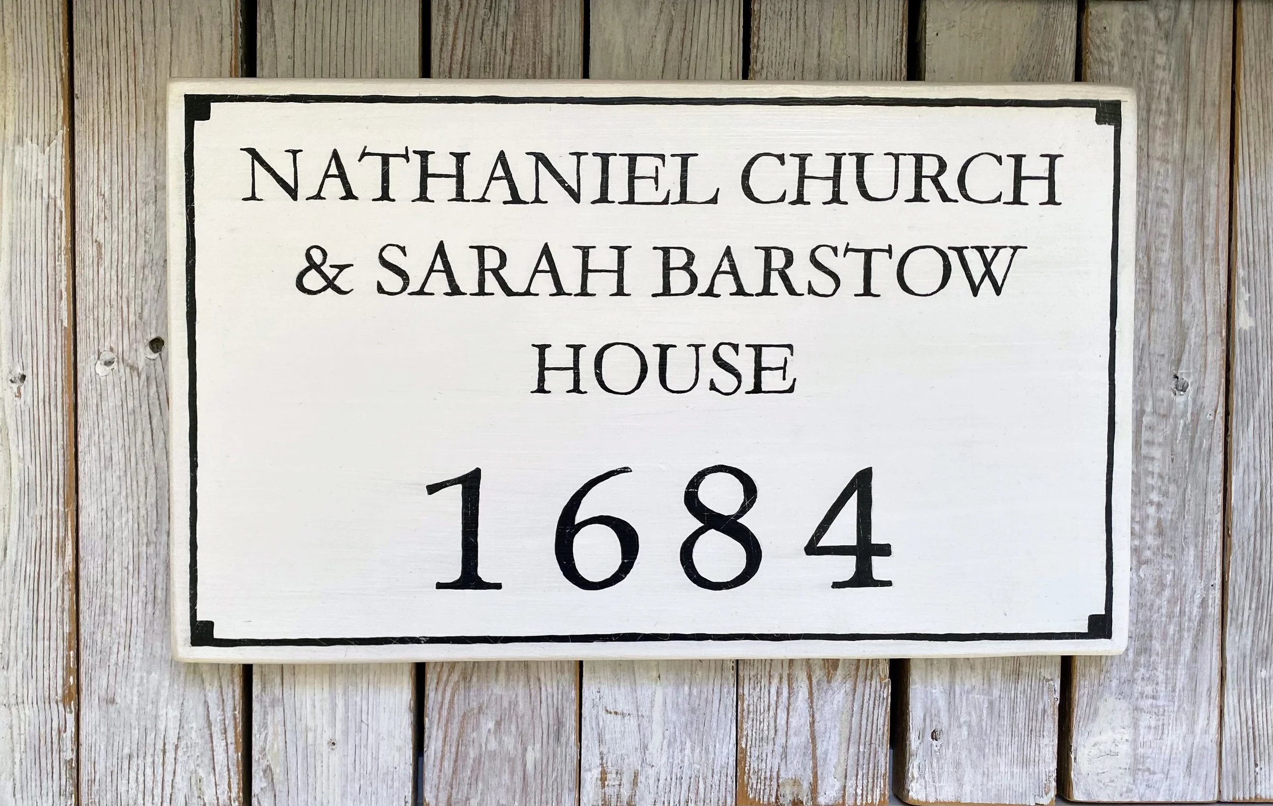 A wooden sign with black lettering reads, 'NATHANIEL CHURCH & SARAH BARSTOW HOUSE 1684,' mounted on a weathered wooden wall.