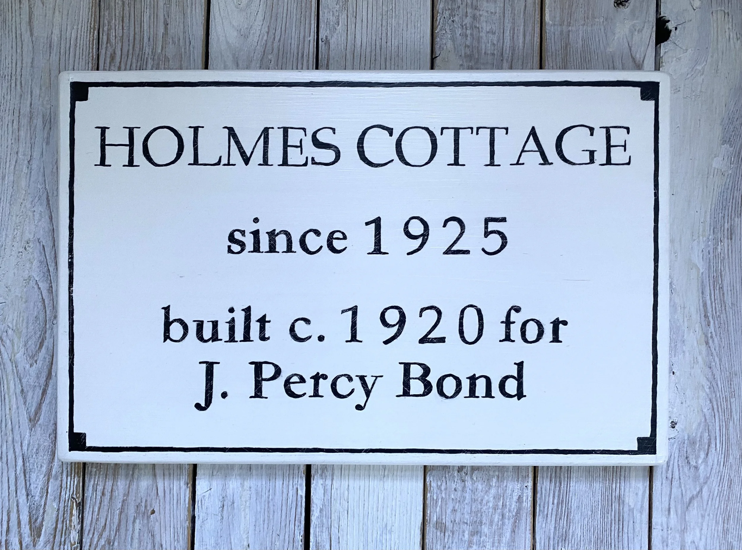 White wooden sign with black border and text reading 'Holmes Cottage since 1925 built c.1920 for J. Percy Bond' mounted on gray wooden wall.