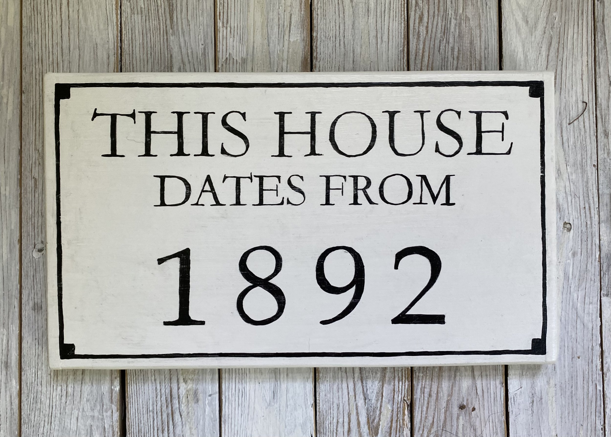 A white wooden sign with black border and lettering that reads 'This house dates from 1892,' placed on a light wood wall.