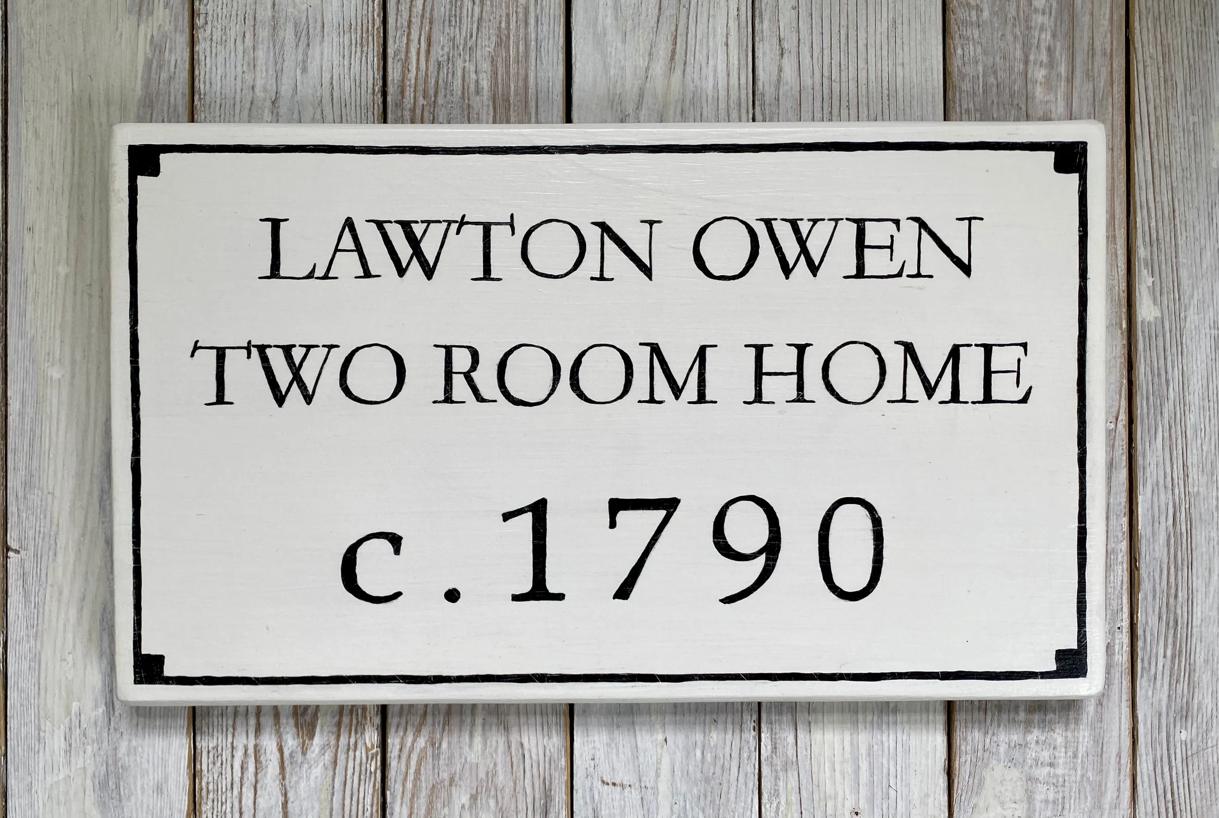 White wooden sign with black border and text reading "Lawton Owen Two Room Home c. 1790" against a light wooden background.