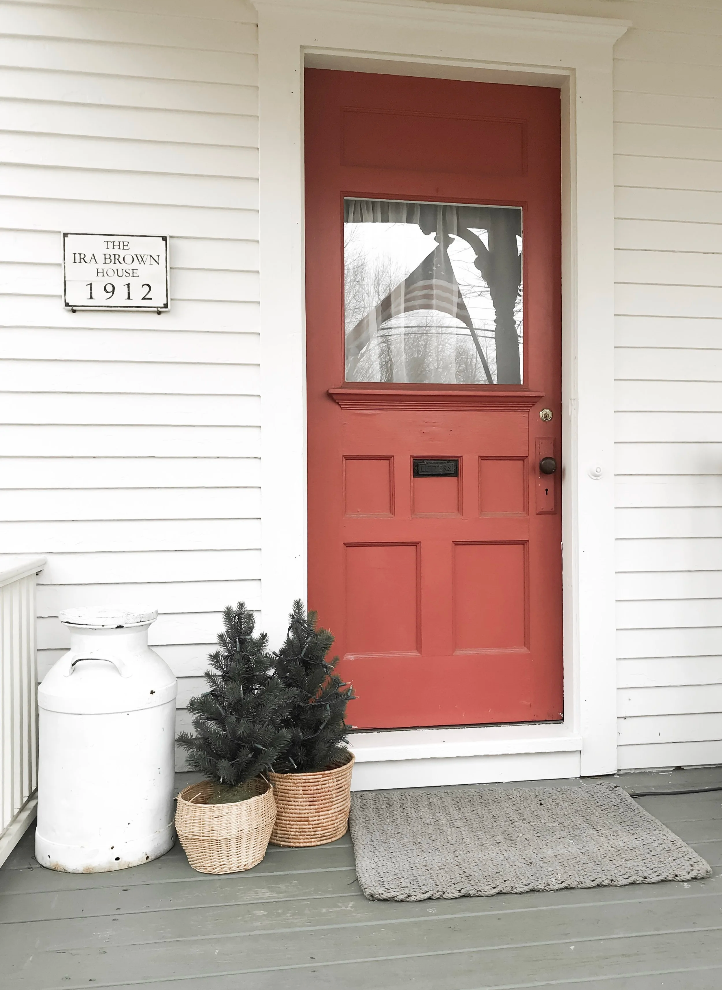 Front door of a house painted red with a window, next to a white wall with a house plaque reading "The Ira Brown House 1912," a white milk can, two baskets with small evergreen trees, and a gray doormat on a wooden porch.
