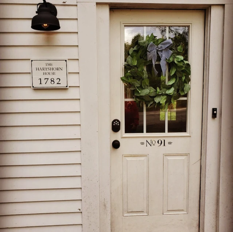 White door decorated with a green wreath and blue-striped ribbon, labeled No 91, on a building with a house number plaque reading 1782 and the name The Hartshorn House.