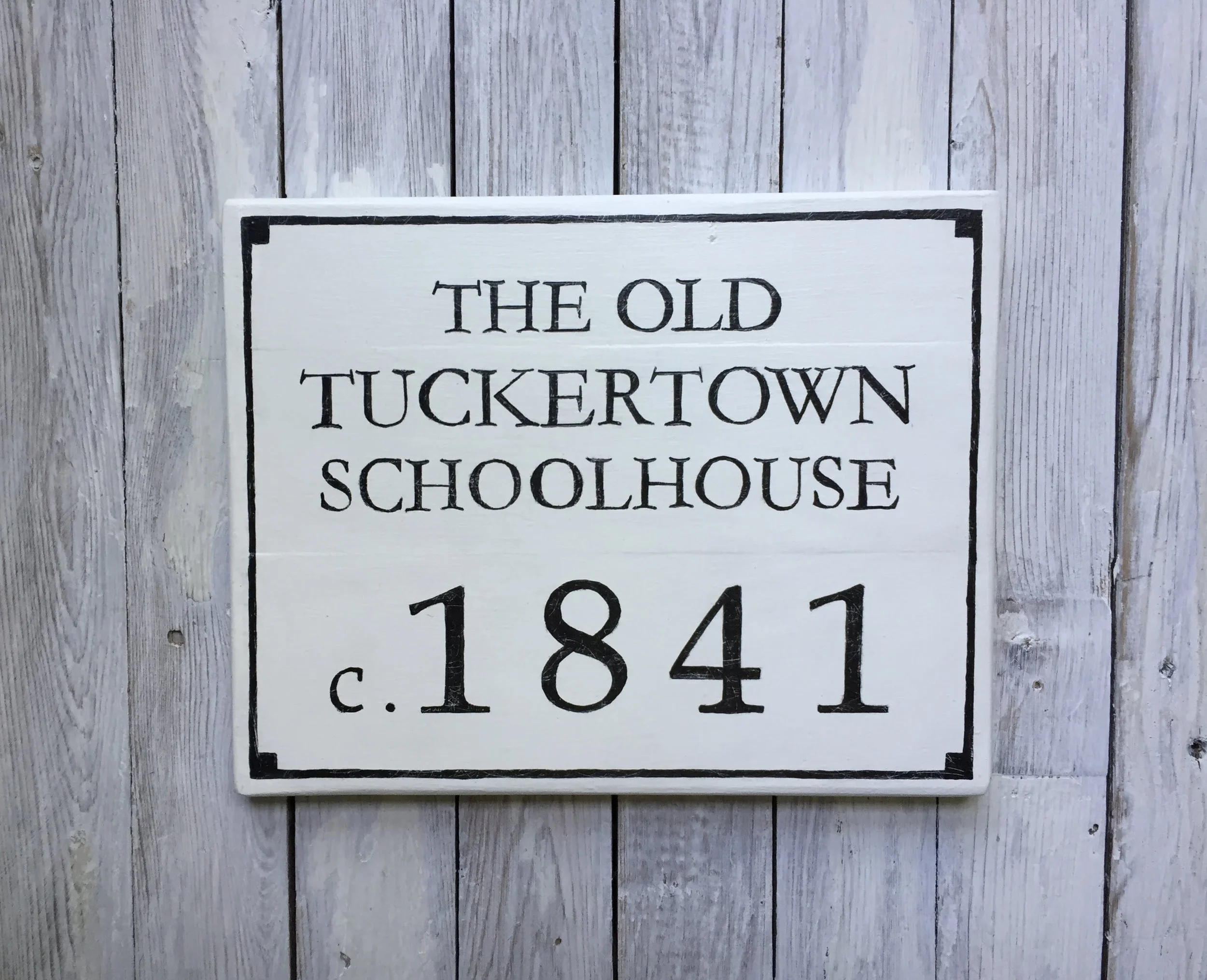 A white wooden sign with black lettering that reads 'The Old Tuckertown Schoolhouse c. 1841', 